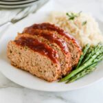 Slices of ground chicken meatloaf topped with a glossy tomato-based glaze, served with roasted asparagus and a side of rice on a white plate.