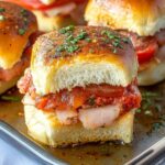 A close-up of an Italian slider made with soft golden-brown buns, layers of deli meat, marinara sauce, and melted cheese, garnished with herbs and baked to perfection.