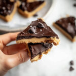 Close-up of a hand holding two stacked pieces of healthy cookie dough bark with a chocolate topping and chocolate chips on top.