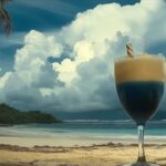 A tropical cocktail with a creamy top and deep blue base served in a glass with a straw, set against a dramatic Caribbean beach backdrop with towering white clouds and turquoise ocean waves.