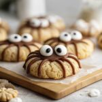 Cute spider-shaped Halloween cookies decorated with chocolate legs and big candy eyes on a wooden board.