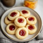 A plate of golden thumbprint cookies filled with red fruit jam, arranged neatly on a neutral-toned ceramic plate.