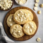 Soft and chewy white chocolate chip cookies stacked on a wooden plate with extra white chocolate chips scattered around.
