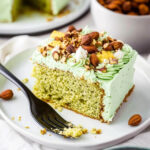 A slice of green pistachio pudding cake topped with fluffy green frosting and crushed almonds, served on a white plate with a black fork.