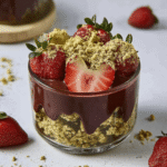 Glass dessert cup filled with crushed pistachios, rich chocolate ganache, and fresh strawberries, topped with gold dust and a halved strawberry.