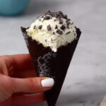 Hand holding a dark chocolate cone filled with creamy vanilla ice cream topped with chocolate shavings