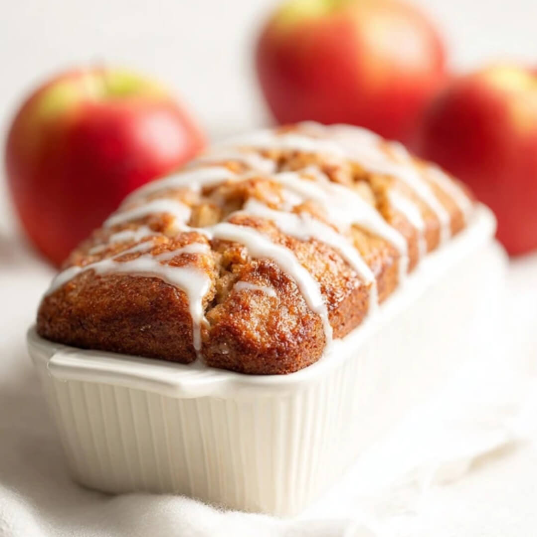 Freshly baked apple cinnamon crumble bread with icing drizzle in a white loaf pan, surrounded by red apples.