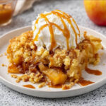 Close-up of caramel apple dump cake topped with whipped cream and drizzled caramel sauce on a plate.
