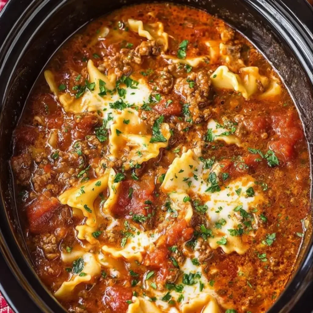 Close-up of Crockpot Lasagna Soup in a black slow cooker, showing crumbled ground meat, broken lasagna noodles, rich red tomato broth, and dollops of melted cheese garnished with fresh parsley.