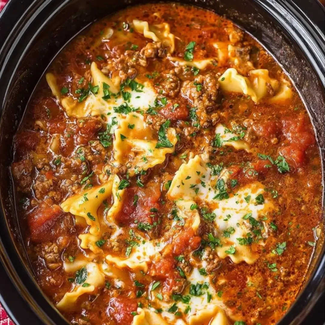 Close-up of Crockpot Lasagna Soup in a black slow cooker, showing crumbled ground meat, broken lasagna noodles, rich red tomato broth, and dollops of melted cheese garnished with fresh parsley.