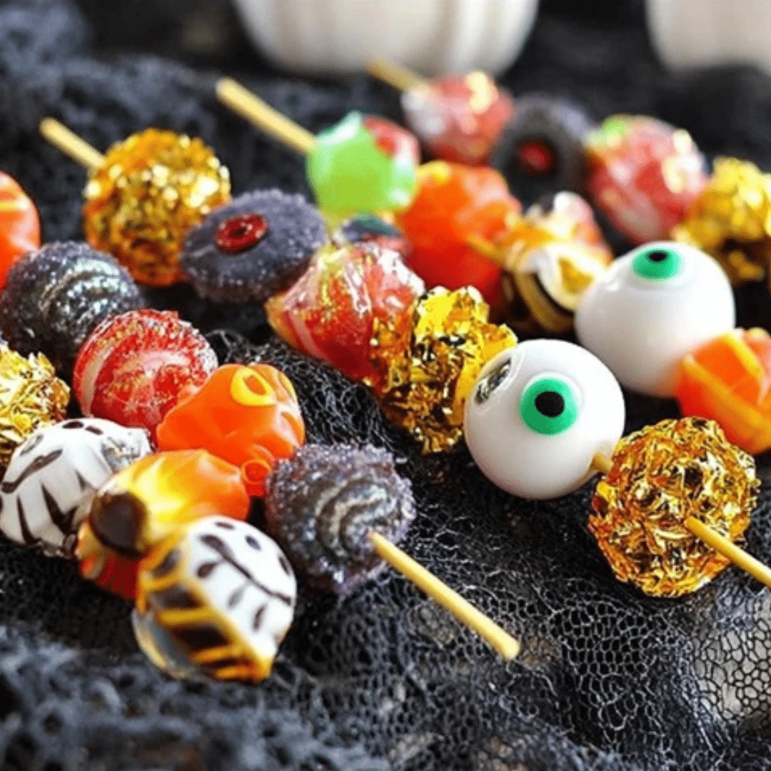 Colorful Halloween candy skewers with gummies, marshmallows, and candy eyeballs on bamboo sticks, displayed on black lace.