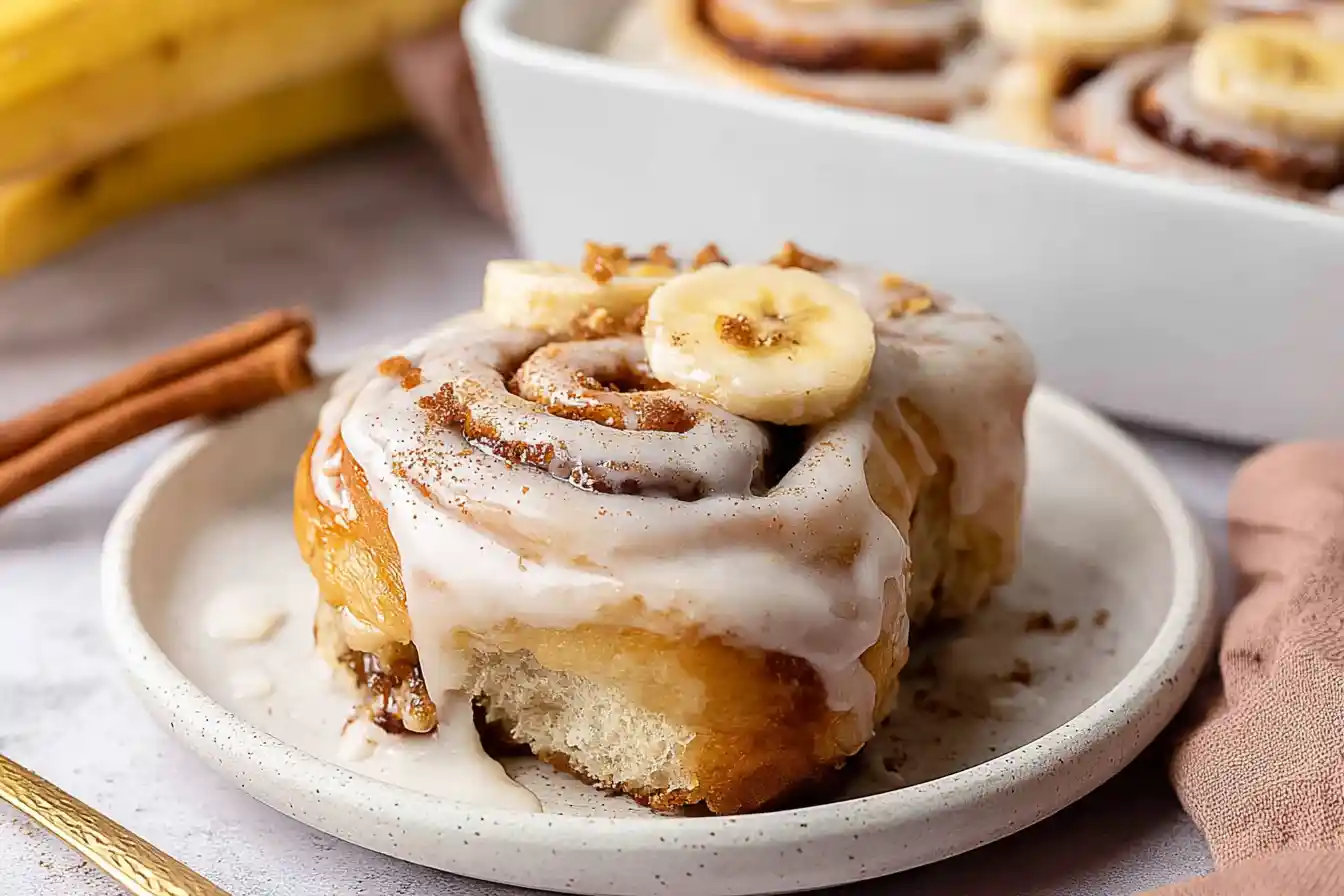 A close-up of freshly baked Banana Bread Cinnamon Rolls, generously drizzled with white cream cheese frosting, on a cooling rack.