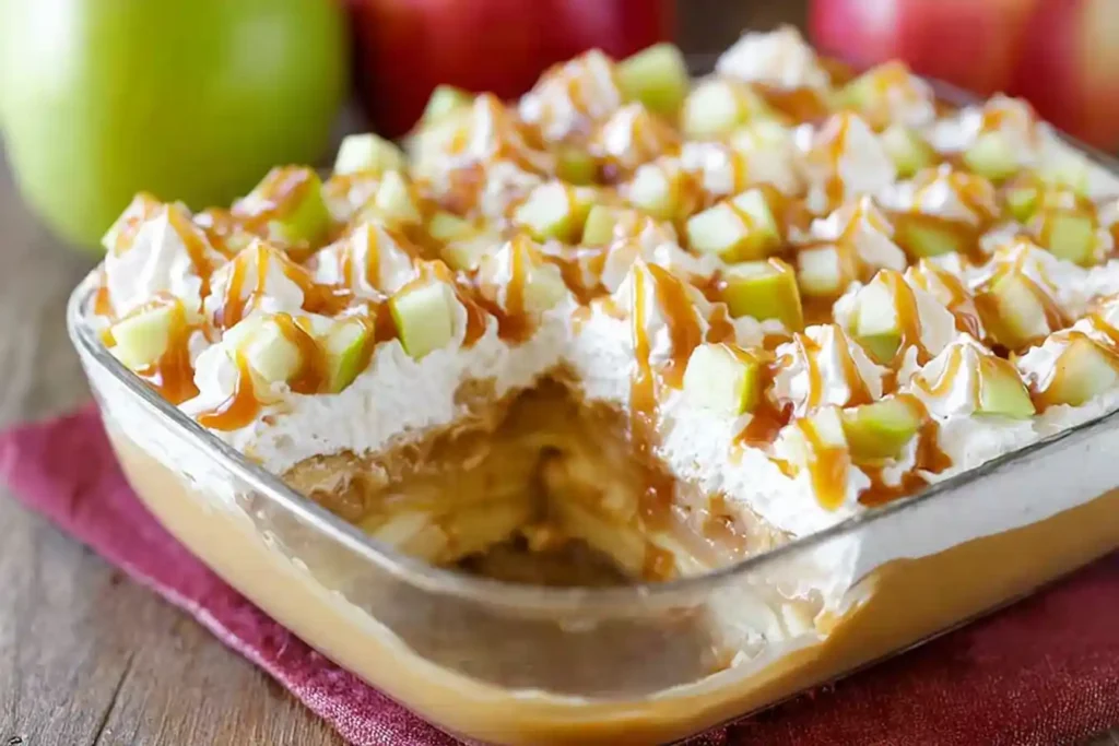 A clear glass bowl filled with layers of creamy Caramel Apple Pudding Dessert, topped with a dollop of whipped cream and a drizzle of caramel sauce.