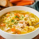 A steaming bowl of homemade chicken noodle soup with fresh herbs and a spoon resting on the rim.
