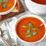 A rustic white bowl of creamy tomato basil soup, garnished with a swirl of cream and fresh basil leaves, sits on a wooden surface.