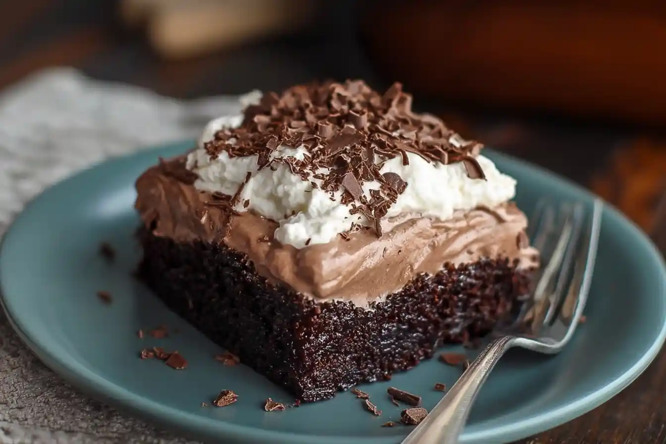 A moist slice of Bailey's Chocolate Poke Cake topped with a creamy white frosting and chocolate shavings is displayed on a white plate.