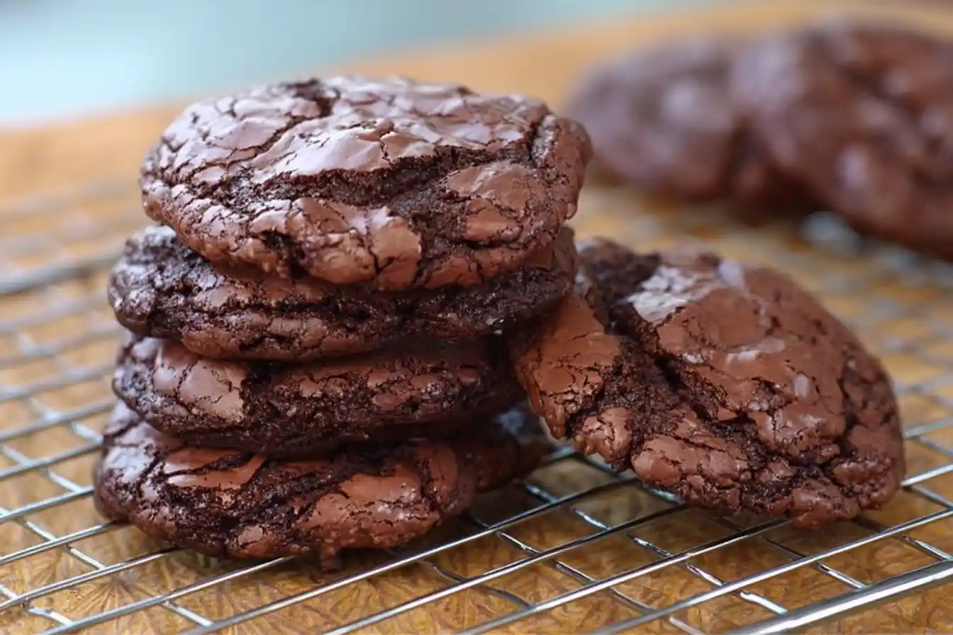 A close-up shot of several freshly baked, crinkled-top brownie cookies stacked on a light surface.