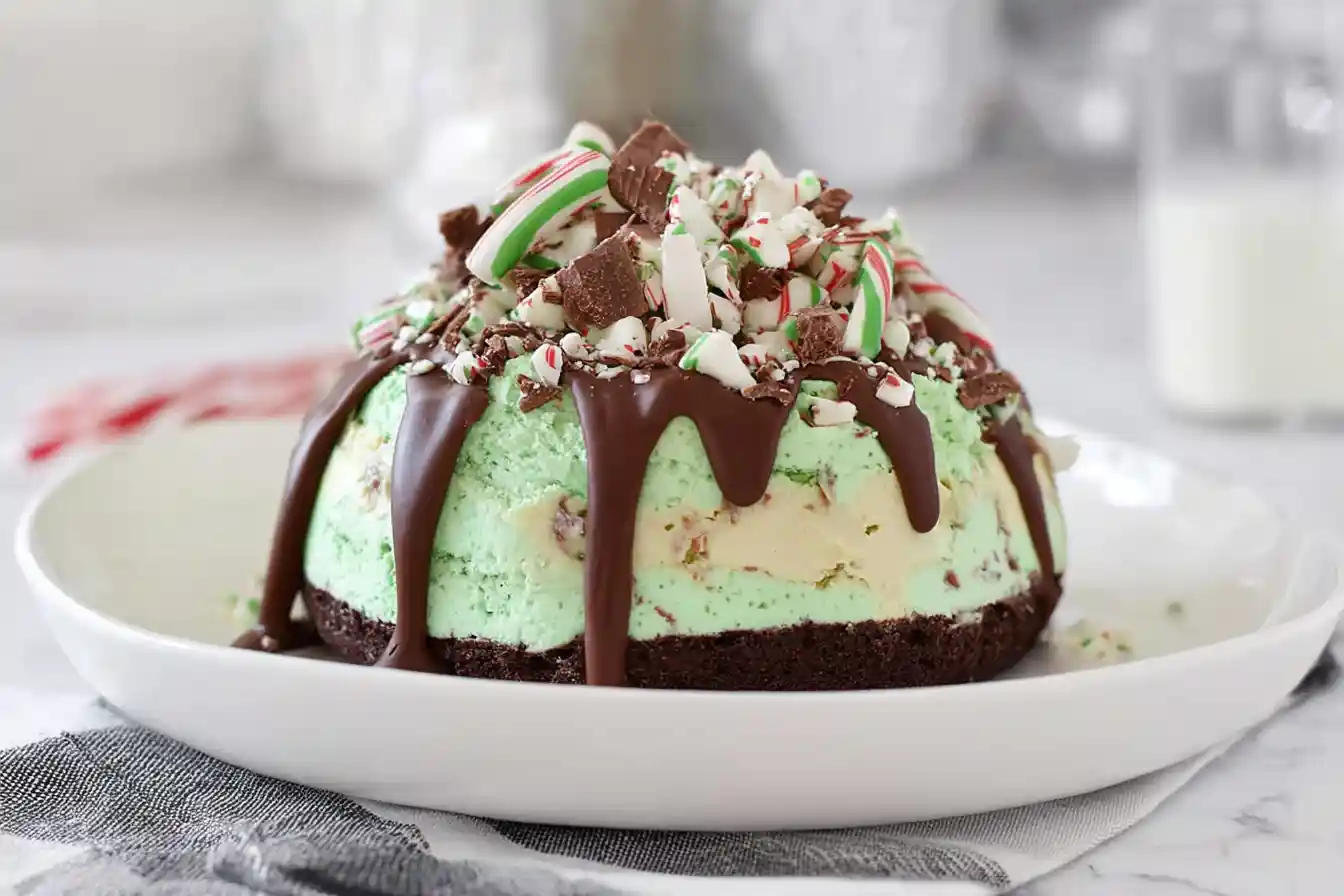 A festive slice of Christmas Peppermint Ice Cream Cake is presented on a white plate, garnished with whipped cream, chocolate shavings, and a mini candy cane.