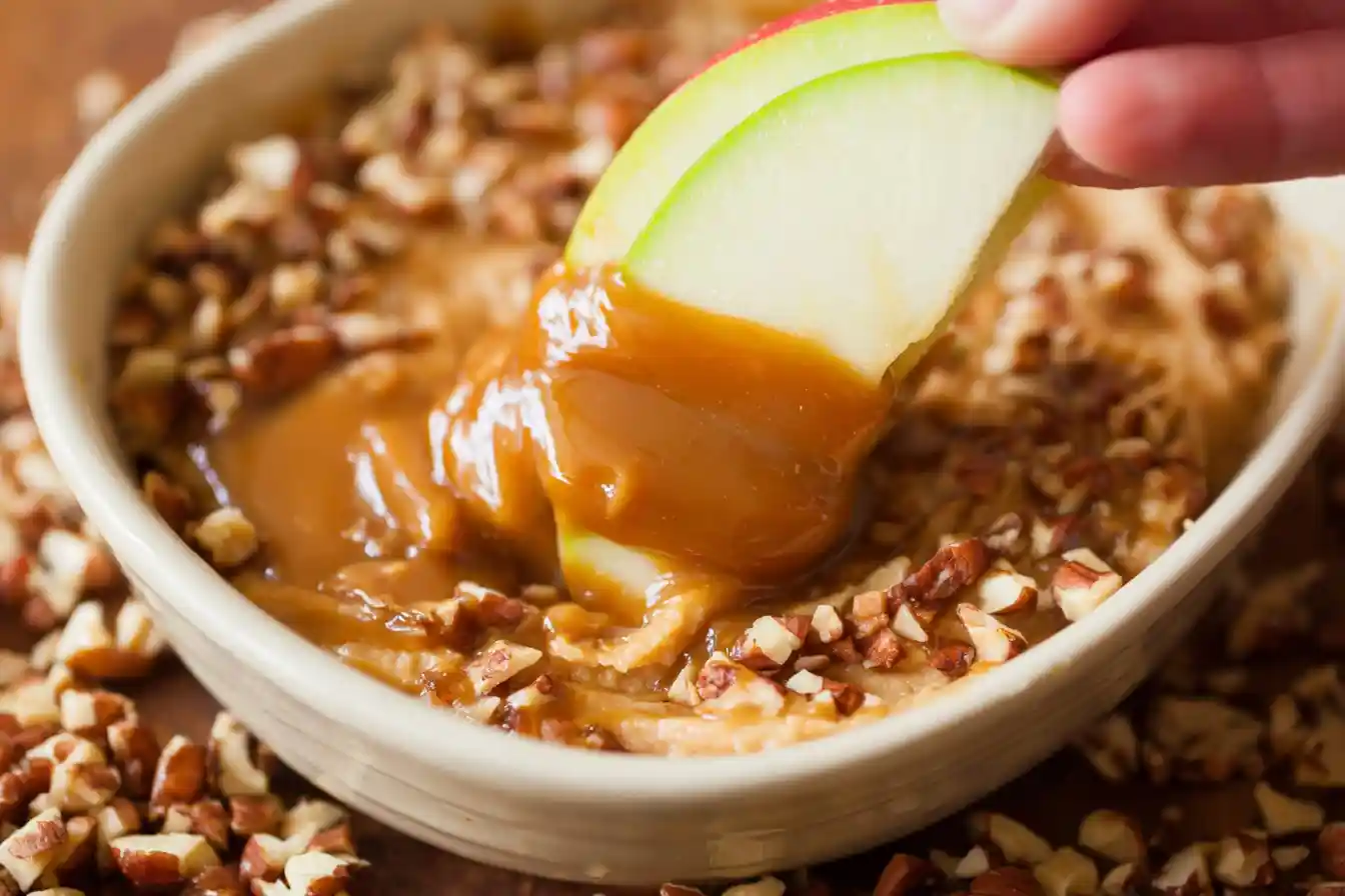 A white bowl filled with creamy Cream Cheese Caramel Apple Dip, topped with a generous swirl of caramel sauce and chopped nuts, surrounded by fresh apple slices ready for dipping.