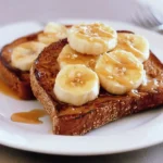 A beautifully arranged slice of Protein Maple Banana Toast is shown, garnished with banana slices, a generous drizzle of maple syrup, and a sprinkle of chopped nuts on a white plate.