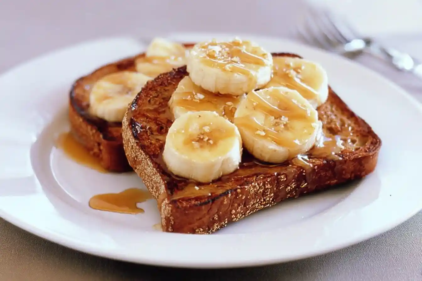 A beautifully arranged slice of Protein Maple Banana Toast is shown, garnished with banana slices, a generous drizzle of maple syrup, and a sprinkle of chopped nuts on a white plate.