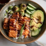 A close-up view of a beautifully prepared Teriyaki Salmon Bowl, featuring a glazed salmon fillet, white rice, edamame, avocado slices, and cherry tomatoes.