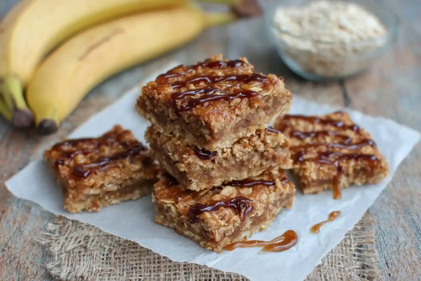 A stack of chewy banana oatmeal bars drizzled with caramel on parchment paper, with fresh bananas and a bowl of oats behind them.