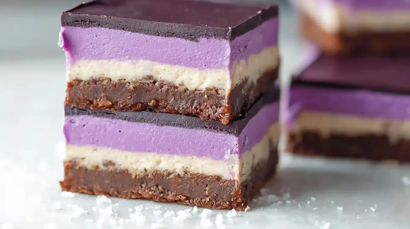 A close-up of two stacked Ube Nanaimo Bars, showing their distinct layers of coconut crust, custard, purple ube, and chocolate.