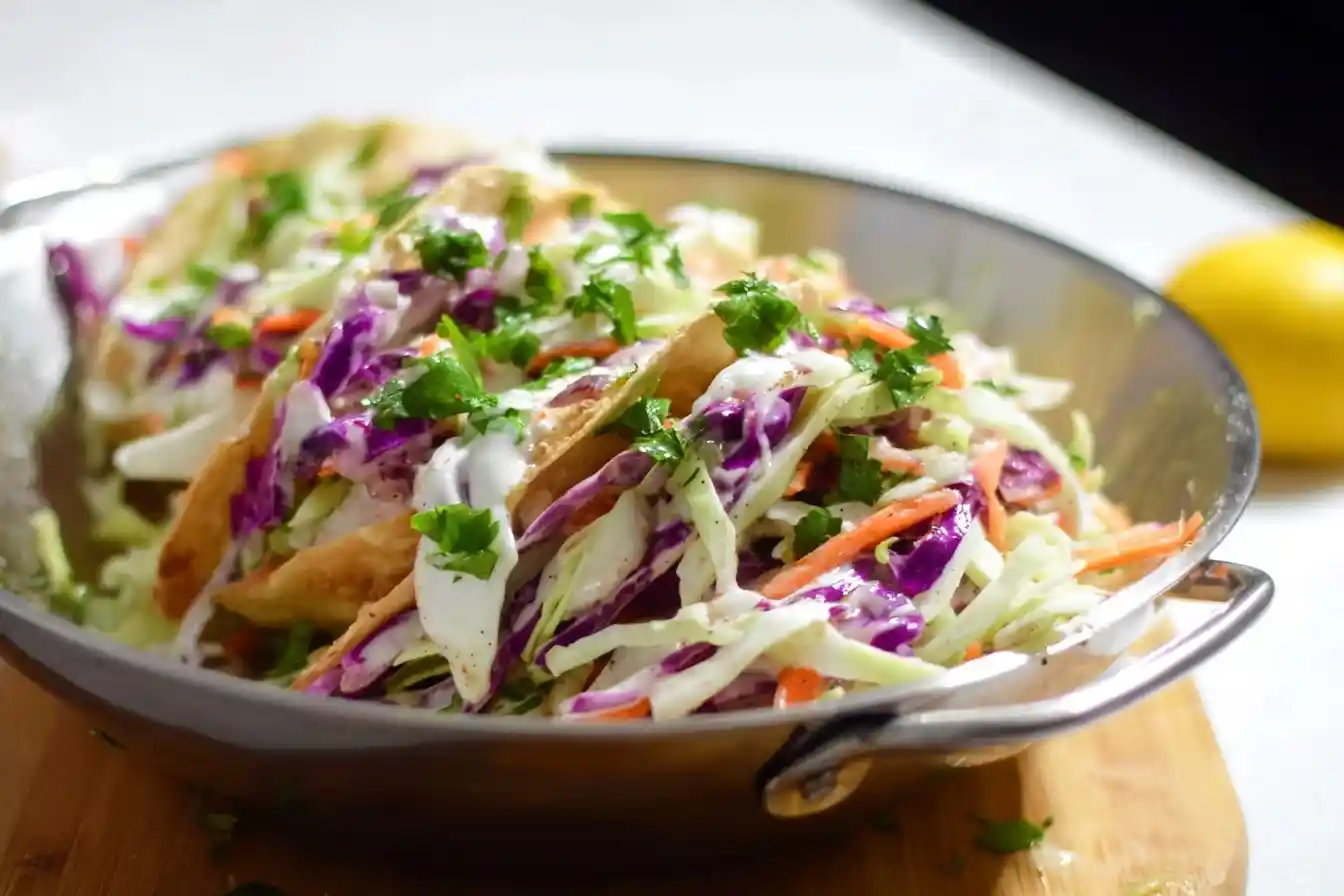 A close-up of several Crispy Fish Tacos in a metal bowl, topped with colorful slaw, creamy sauce, and fresh cilantro.