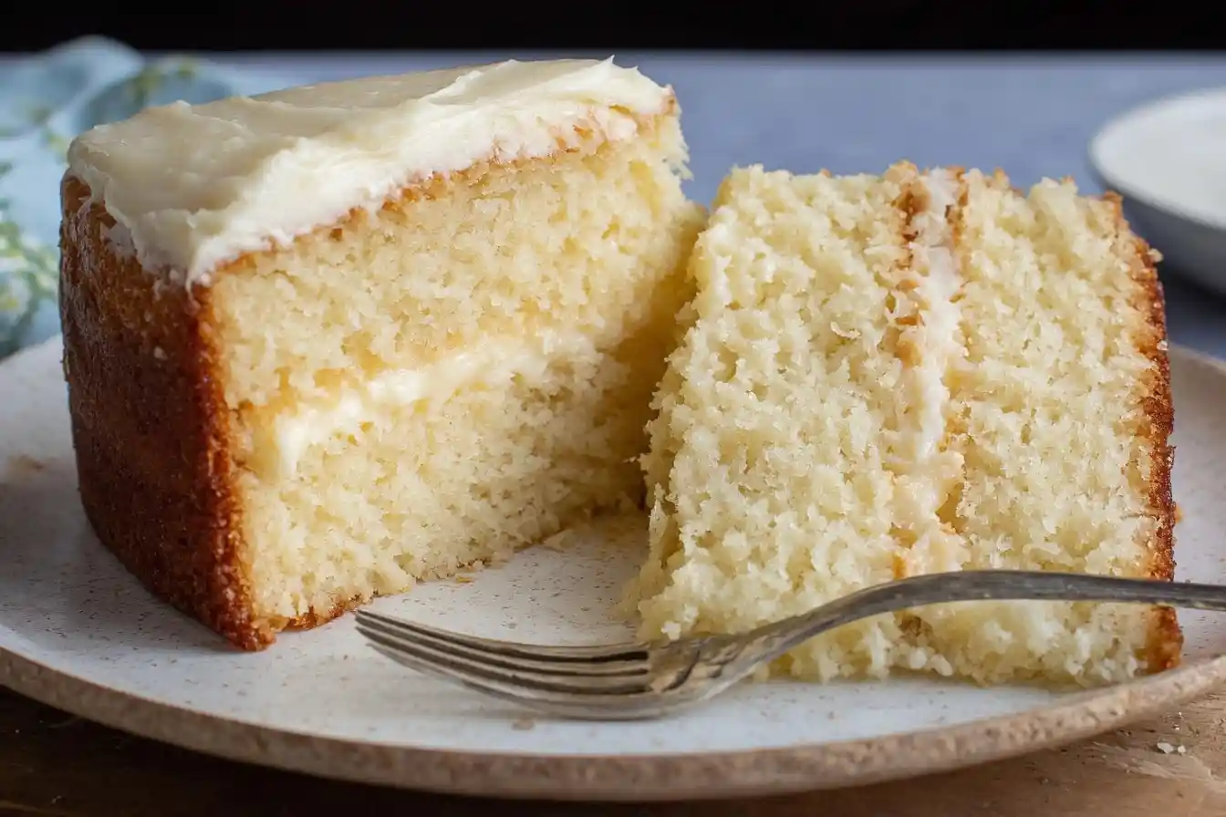 A slice of a two-layer vanilla cake with white frosting sits on a speckled plate next to a fork, showing its moist crumb.