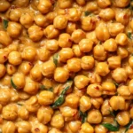 A close-up overhead shot of a creamy Chickpea Curry with spinach and chili flakes, with a wooden spoon stirring the dish.