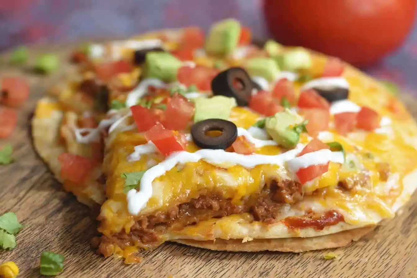 A close-up of a homemade Mexican Pizza on a wooden board, topped with cheese, tomatoes, olives, and avocado.