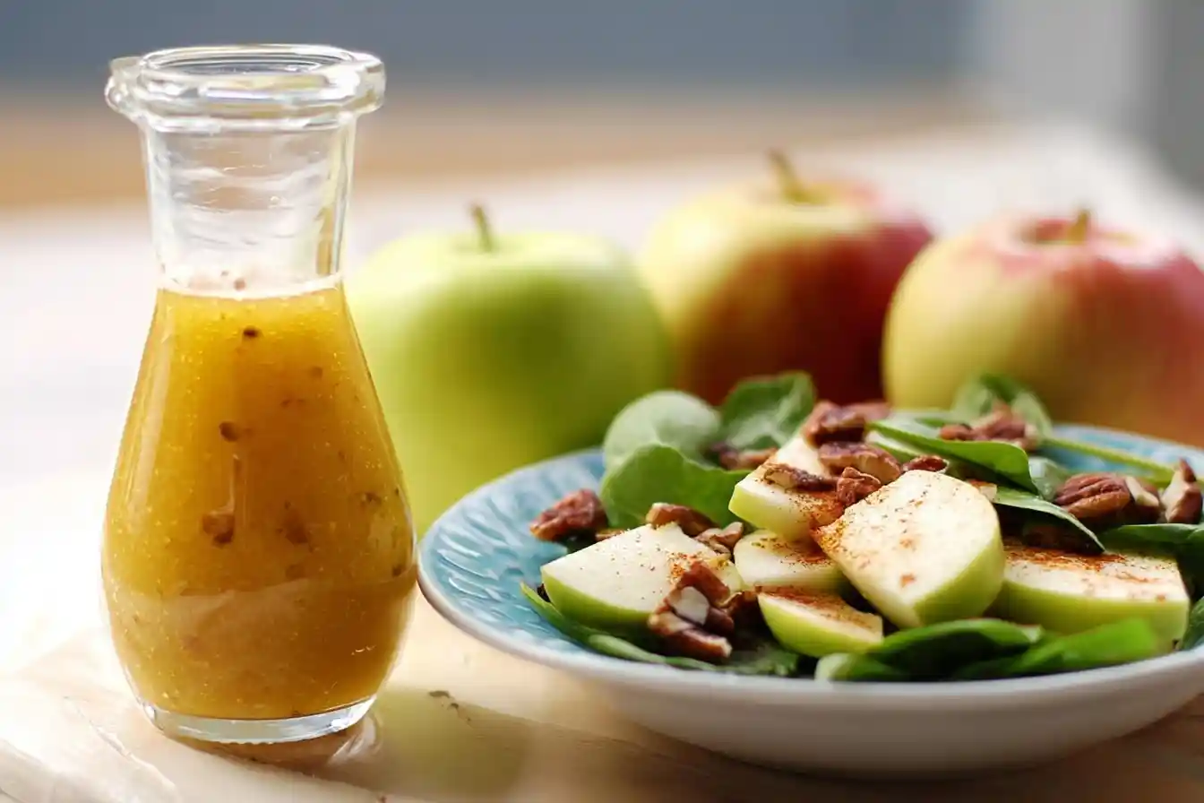 Glass cruet of golden Apple Cider Vinaigrette sitting next to a fresh spinach salad with sliced apples and pecans.