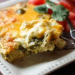 Close-up of a cheesy Chile Relleno Casserole slice served on a white plate with fresh tomatoes and cilantro.