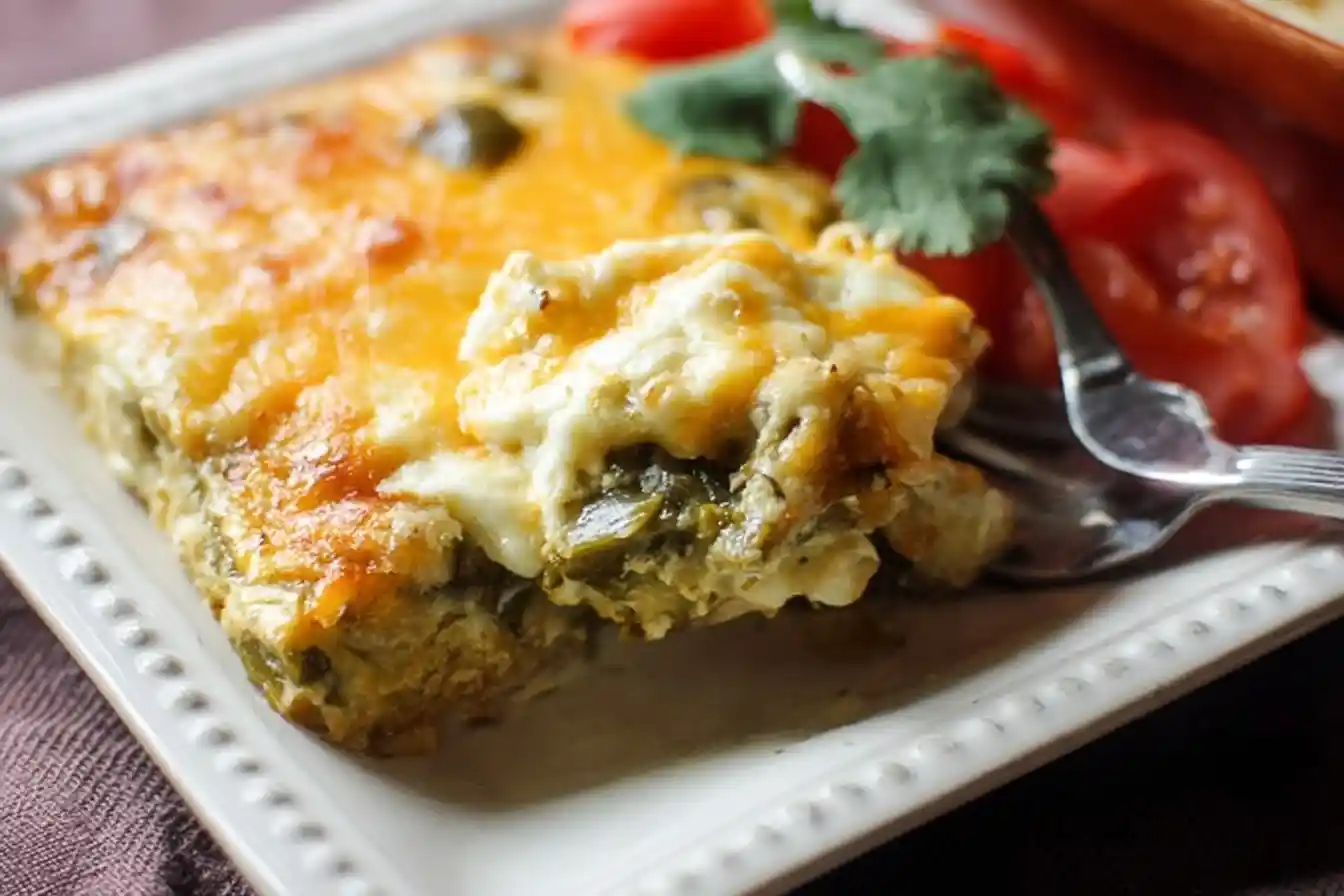 Close-up of a cheesy Chile Relleno Casserole slice served on a white plate with fresh tomatoes and cilantro.