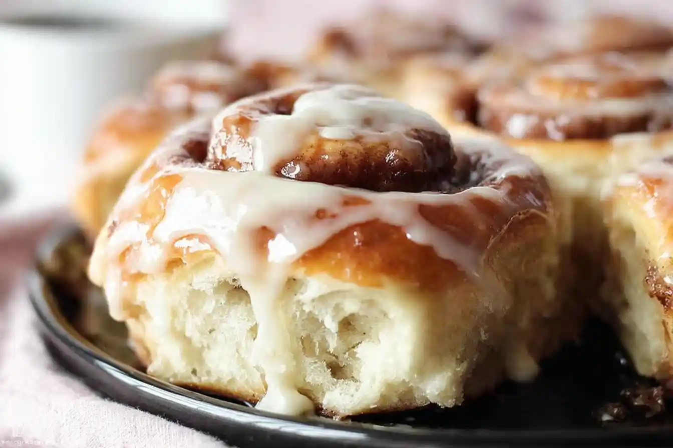 A close-up shot of a fluffy, warm cinnamon roll on a black plate, with sweet white icing dripping down the side.