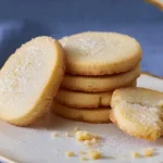 Stack of buttery shortbread cookies sprinkled with sugar on a white plate with a broken cookie in front.