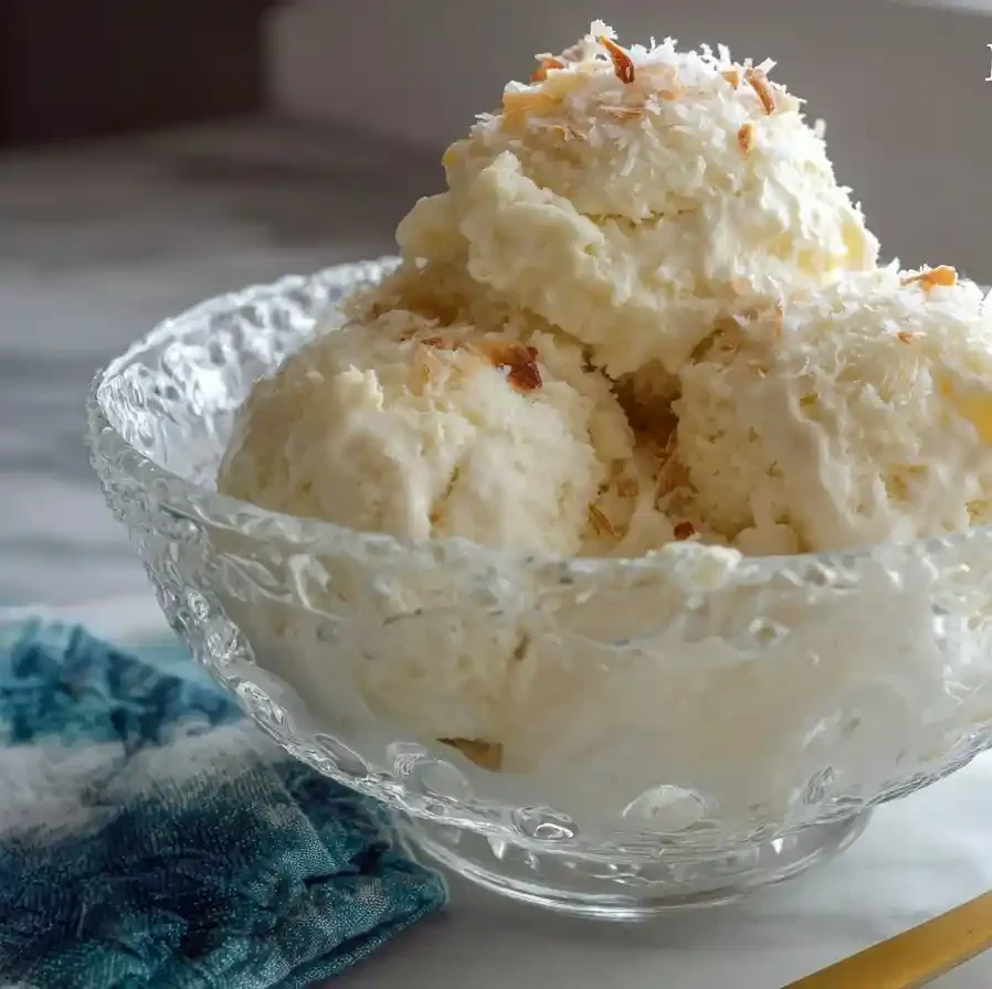 A clear glass bowl filled with scoops of homemade coconut ice cream and topped with toasted coconut flakes, next to a gold spoon.