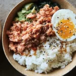 Top-down view of a salmon rice bowl topped with flaked salmon, avocado, seaweed, and a soft-boiled egg.