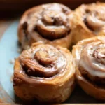 A close-up of warm, glazed cinnamon rolls on a blue plate beside a speckled mug of coffee.