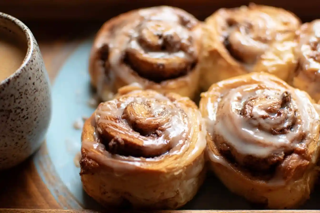 A close-up of warm, glazed cinnamon rolls on a blue plate beside a speckled mug of coffee.