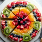 A beautifully sliced fruit pizza on a white platter, topped with cream cheese frosting, fresh berries, kiwi, and oranges.
