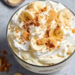 A glass bowl filled with creamy banana pudding, topped with whipped cream, fresh banana slices, and crushed wafers.