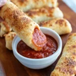 A hand dipping crispy baked pepperoni pizza rolls into a white bowl of red dipping sauce on a wooden board.