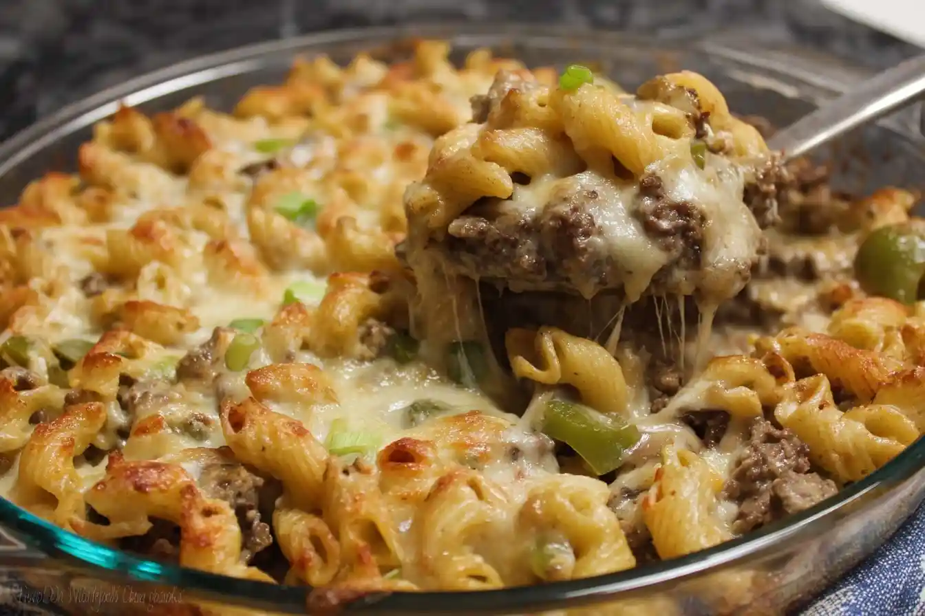 A serving spoon lifting a scoop of cheesy Philly Cheesesteak Pasta with ground beef and peppers from a glass dish.