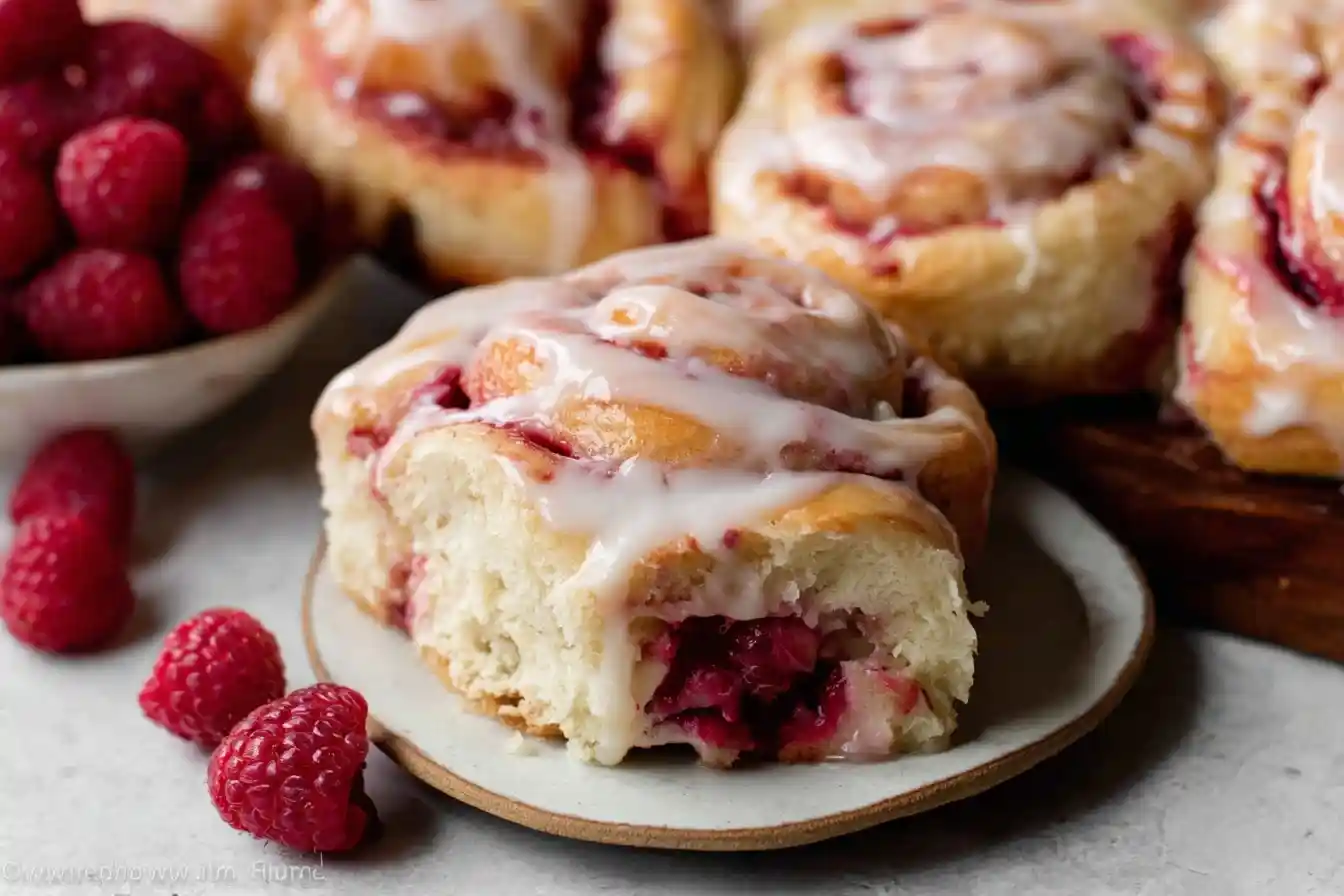 A close-up of gooey raspberry sweet rolls topped with sweet icing, served on a plate with fresh raspberries.