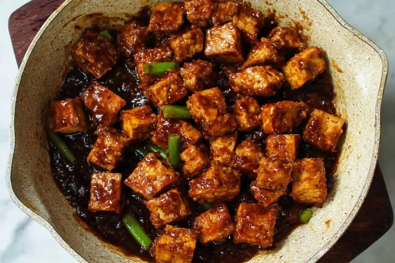 Overhead view of crispy General Tso's Tofu simmering in a savory dark sauce with green onions in a skillet.
