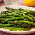A white plate heaped with glossy sauteed asparagus spears topped with minced garlic, next to a fresh lemon.