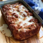 Freshly baked cinnamon roll loaf covered in sweet white icing, sitting in a metal pan on a wooden board.