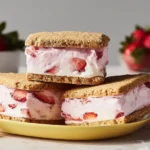 A stack of three homemade strawberry ice cream sandwiches on a yellow plate with fresh strawberries in the background.